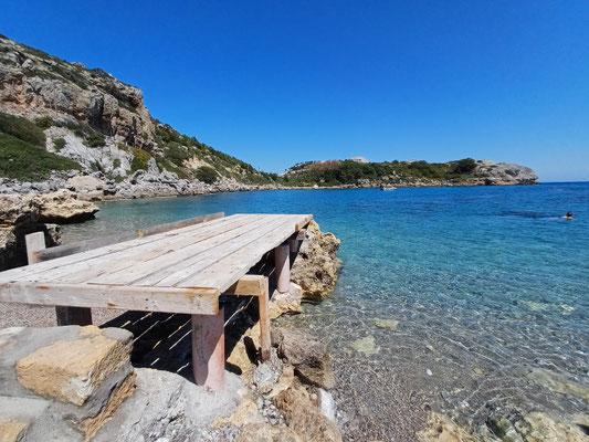 Auf der linken Seite des Fotos ist rangezoomt ein Steg zu sehen, der ins Meer führt. Im Vordergrund ist ein Kiesstrand zu sehen, dahinter das türkisblaue Meer mit Küste.