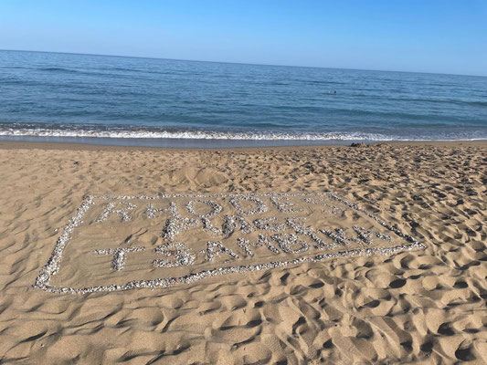 Auf dem Foto sieht man einen Schriftzug "Rhodes Tsambika", der aus Steinen am gelegt wurde . Im Hintergrund ist das Meer zu sehen.