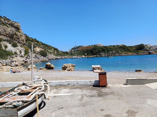 Auf der linken Seite des Fotos ist ein Holzboot zu sehen, dahinter sieht man den Kiesstrand und das Meer mit der Küste.