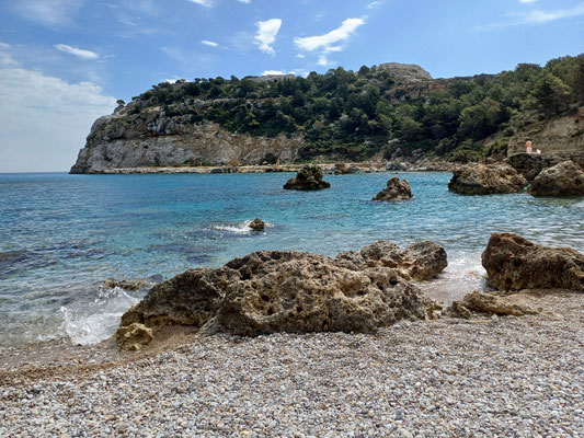 Im vorderen Bereich des Fotos ist der Kiesstrand zu sehen, dahinter ein größerer Felsen sowie das türkises Meerwasser und weitere Felsen im Hintergrund.