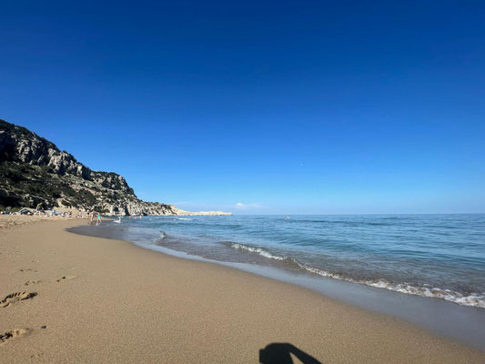 Im Vordergrund ist der goldene Sandstrand zu sehen. Im rechten Teil ist das Meer und der blaue Himmel zu sehen. Auf der linke Seite ist der Beginn der Küste zu erkennen.