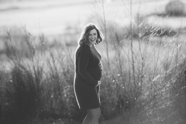 Werdende Mutter im figurbetonten Strickkleid lacht ausgelassen beim Babybauch-Shooting in der Natur.