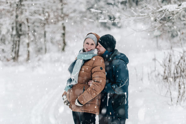  Glückliche werdende Eltern beim Winter-Babybauch-Shooting: Sie lachen eng aneinandergekuschelt im Schnee