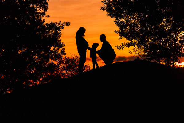 Schwangere beim emotionalen Familienshooting mit Partner und Geschwisterkind im Sonnenuntergang.