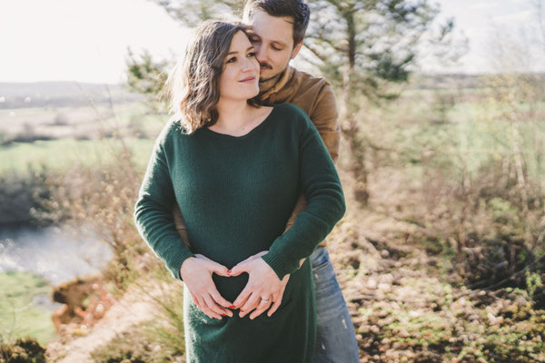 Paar formt mit den Händen ein Herz auf dem Babybauch beim Schwangerschaftsshooting in der Natur.