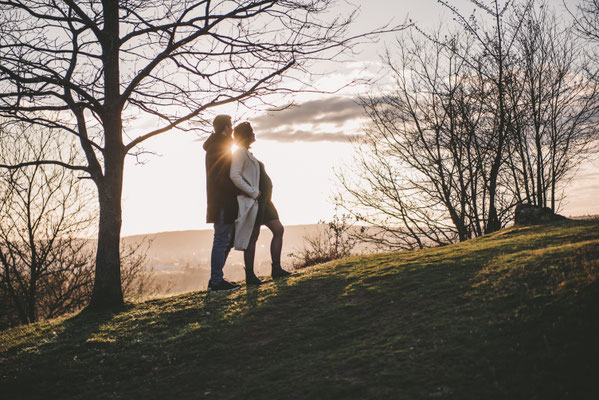 Natürliches Babybauch-Shooting im Herbst: Werdende Eltern stehen bei Sonnenuntergang auf einem Hügel.