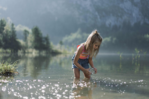 Kind spielt am flachen Ufer des Traunsees im klaren Wasser mit Berglandschaft im Hintergrund