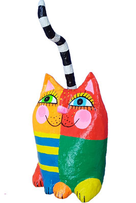 Handgefertigte Pappmaché-Katzenfigur in leuchtenden Farben: linker Körperbereich in Gelb mit blauen Streifen, rechter Bereich in Rot und Grün. Die Katze hat rosa Wangen, große blaue Augen mit Wimpern und einen schwarz-weiß gestreiften Schwanz.