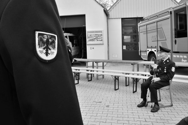 Älterer Herr mit Stock, sitzend, gekleidet in Feuerwehr Uniform. Im Vordergrund ein Bundesadler auf der Uniform eines Kollegen.