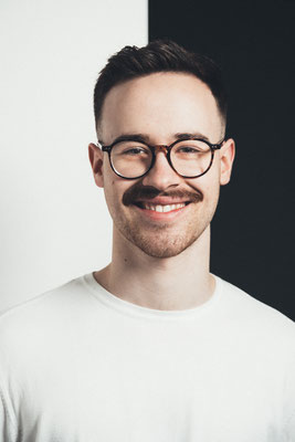 Junger Mann mit Brille und Schnurrbart, lächelnd. Im Fotostudio vor schwarzweißem Hintergrund.