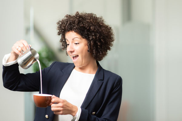 Junge, fröhliche Frau schenkt sich Milch in eine Kaffeetasse ein.