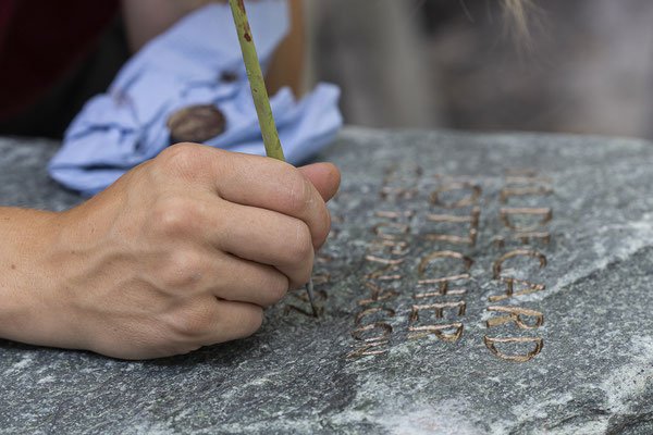 Einer Mitarbeiterin des Steinmetzbetriebes Carl Schütt und Sohn malt mit einem feinen dünnen Pinsel goldene Farbe in die in den Grabstein gravierten Schriftzeichen. Detailaufnahme, es sind nur die Hand mit dem Pinsel uund die Schrift sichtbar.