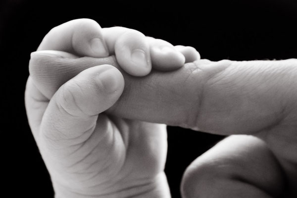 Neugeborene, hier sieht man die Hand eines Babys, welches den Finger ihres Vaters umklammert auf schwarzem Hintergrund