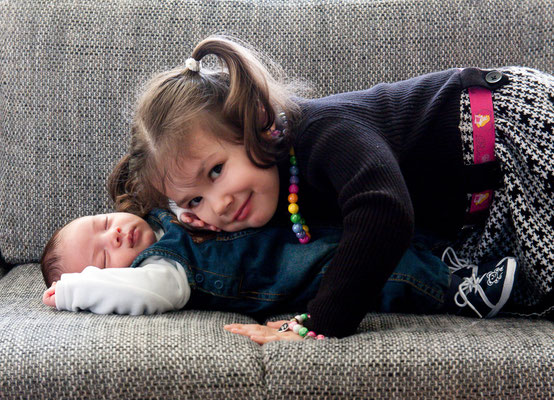 Sie sehen Geschwister auf einem Sofa liegend. Das Baby liegt auf dem Rücken und seine Schwester dient über ihm und schaut in die Kamera.