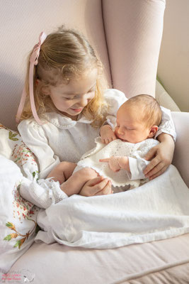 Dies ist das Foto zweier Geschwister in einem Sessel sitzend. Die große Schwester hat das Baby auf dem Arm
