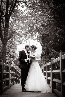 Hochzeitsfotografie, ein Hochzeitspaar im Ahrensburger Schlosspark auf einer Brücke mit einem Herzregenschirm während sie sich küssen