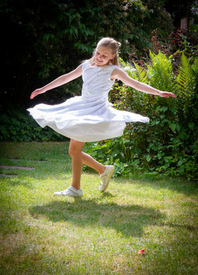 Sie sehen ein Mädchen im Kommunionkleid, welches sich in einem Garten dreht, dass das Kleid fliegt.