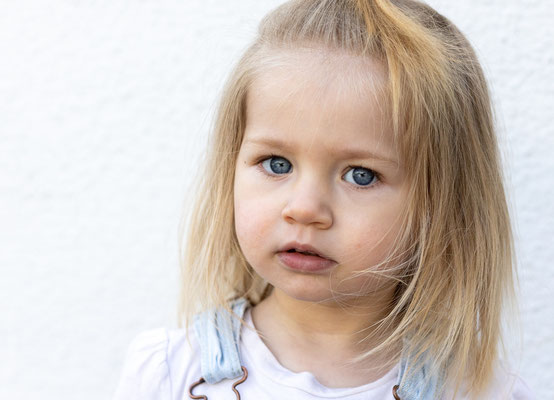Hier sehen Sie ein Kopfportrait eines kleinen Mädchens, seitlich angeschnitten und blonden Harren, dass nachdenklich in die Kamera sieht