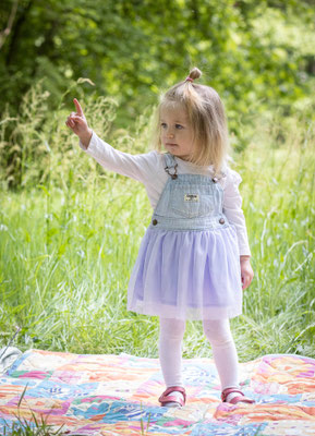 hier sehen Sie ein kleines Mädchen mit Jeanskleid auf einer Picknickdecke stehend im Wald und mit ausgestrecktem Arm zeigend