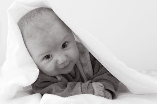 Dieses Bild zeigt ein Baby, welches sich gerade auf die Arme aufstützt mit einem Tuch über dem Kopf in schwarz weiß