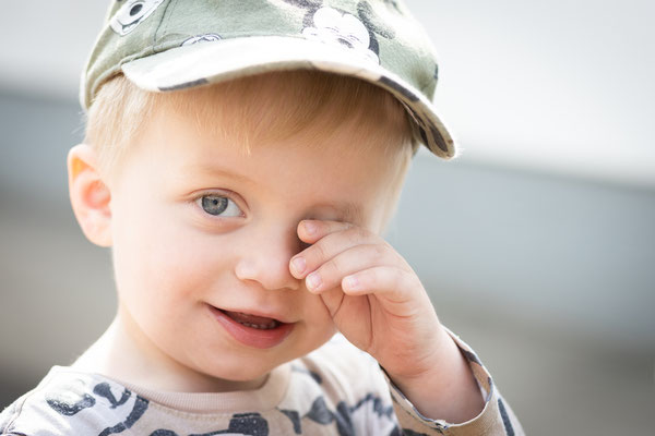 Dieses Foto zeigt einen Kleinkind-Jungen im Kopfportrait, der sich gerade das rechte Auge reibt und niedlich in die Kamera schaut, aus einem Kindergartenshooting beim spielen 