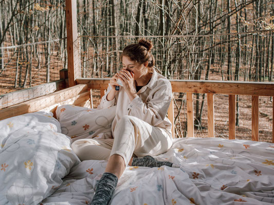 Junge Dame sitzt auf dem Bett und trinkt Kaffeee beim Baumhaus Freiraum.