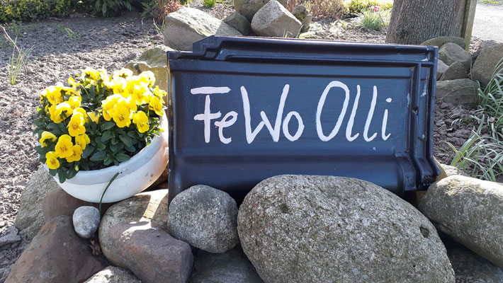 Liebevoll selbstbemalte Dachziegel mit der Aufschrift FeWo Olli als individueller Willkommensgruß im Blumenbeet am Hofeingang.