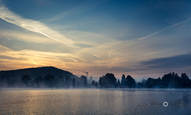 Nebelstimmung bei Sonnenaufgang am Wörthsee (Nebelschwaden über dmr Wasser), im Hintergrund ein Kran, am Himmel von der Sonne angeleuchtete Kondensstreifen und Wolken