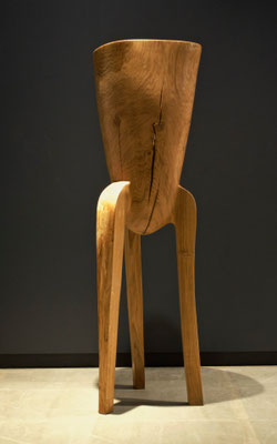 abstract three-legged oak wood sculpture V1386 by Jörg Pietschmann, highlighting a distinctive dark triangular wood inlay and vertical crack, photographed against a dark grey wall.