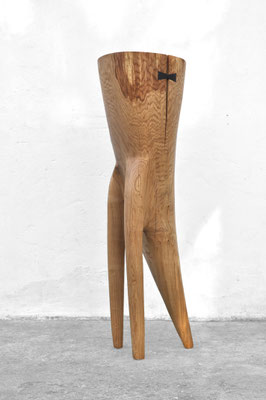 Abstract three-legged oak wood sculpture V1406 by Jörg Pietschmann, featuring a dark bowtie inlay stabilizing a natural wood crack, studio shot on a concrete floor with a white wall background.