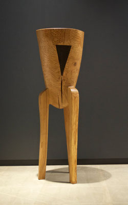 abstract three-legged oak wood sculpture V1386 by Jörg Pietschmann, highlighting a distinctive dark triangular wood inlay and vertical crack, photographed against a dark grey wall.
