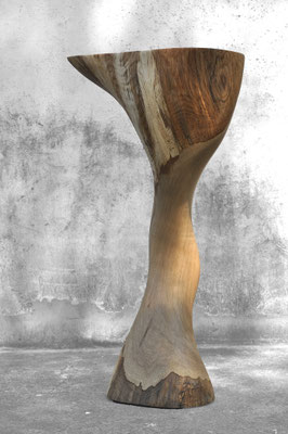 Organic oak wood bowl sculpture V1366 by Jörg Pietschmann, featuring a fluid, curved pedestal and a hollowed top, photographed outdoors against a weathered concrete wall.