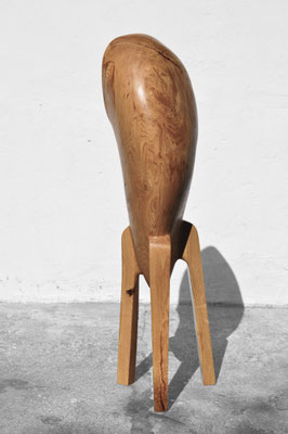 Three-quarter view of the abstract three-legged oak wood sculpture V1376 by Jörg Pietschmann, showing its large hollowed-out interior cavity and smooth organic form, shot on a concrete floor against a white wall.