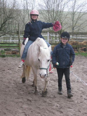 Mädchen reitet auf einem weißen Pony und der Junge führt es. Das Mädchen streckt einen Wollebommel nach rechts.