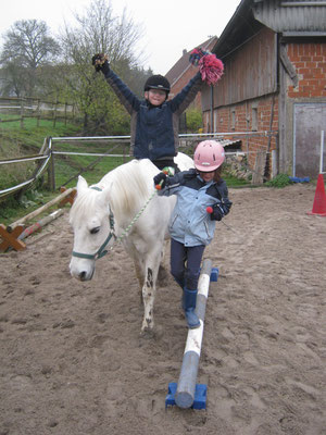 Ein Junge sitzt auf einem weißen Pony und streckt die Arme in die Luft. Ein Mädchen führt das Pony und balanciert über eine Stange.
