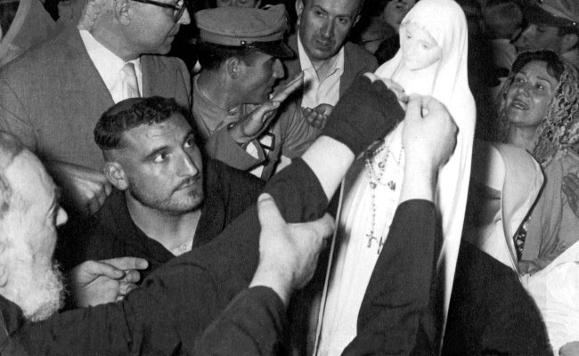 Padre Pio, in quel momento infermo, viene aiutato ad offrire una corona del Rosario, ricevuta in dono dal Gruppo di preghiera di San Casciano Val di Pesa, alla venerata immagine della Madonna Pellegrina