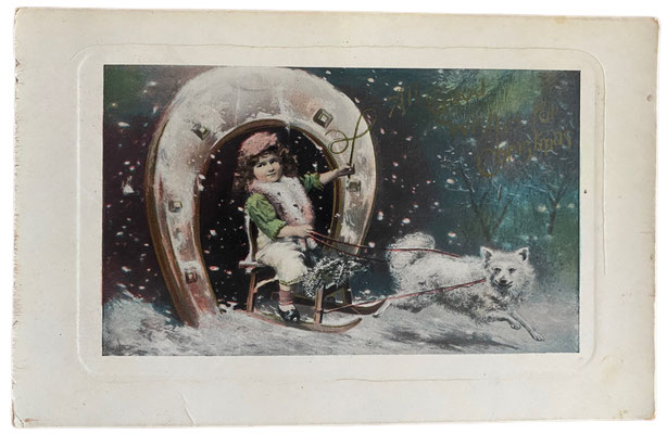 "All good wishes for Christman" in Goldbuchstaben auf einer lackierten Postkarte. Ein weißer Spitz zieht ein Kind in einem Schlitten durch ein hufeisenförmiges Tor.