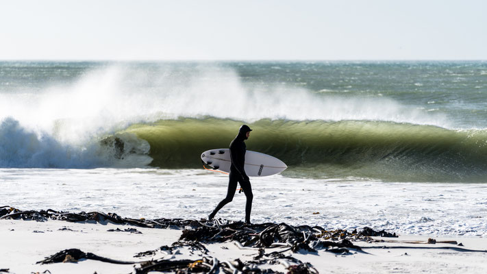 A surfer in a wet suit walking along Kommetjie beach, South Africa