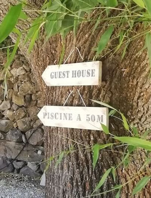 Flèches directionnelles en bois peint en blanc accrochées à un arbre indiquant "piscine" et "guest house" pour un gîte