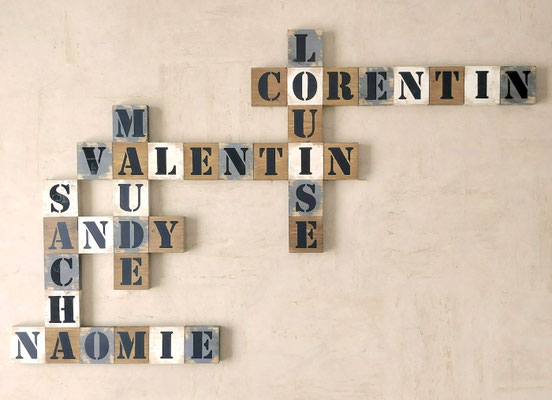 Scrabble mural en bois, gris et blanc, personnalisé avec prénoms de la famille peints en noir, accroché sur un mur couleur sable