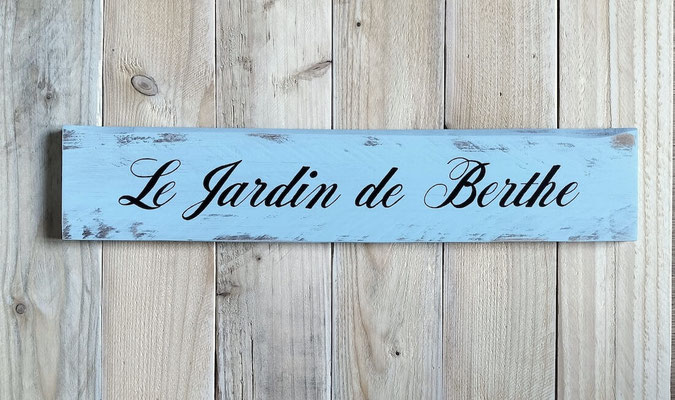 Pancarte peinte en bleu clair posée sur un fond bois clair indiquant "Jardin de Berthe" en noir