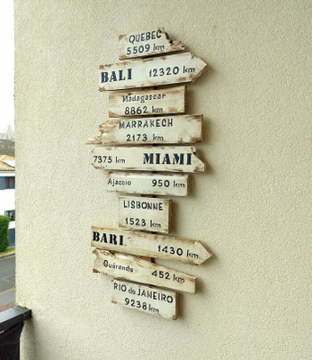 Accroché sur mur clair en extérieur, panneau de direction bois blanc vieilli personnalisé avec des villes comme Marrakech, Bali ou Miami