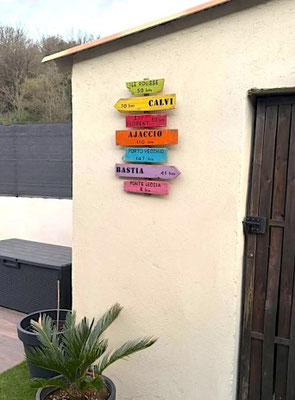 Panneau directionnel bois personnalisé, couleurs vives et destinations de Corse peintes en noir avec des km accroché en extérieur à côté d'une plante verte