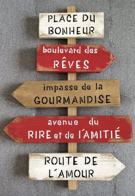 Panneau de direction rouge et blanc posé sur un mur gris personnalisé avec des phrases comme "place du bonheur" ou "avenue du rire et de l'amitié"