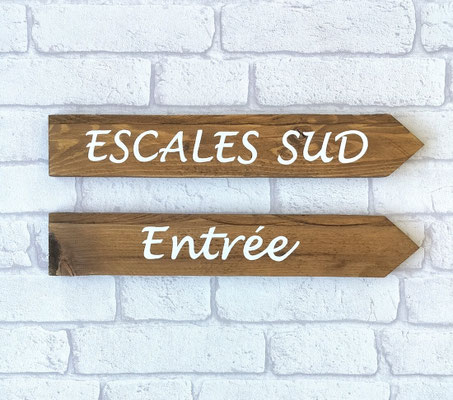 Flèches en bois indiquant l'entrée d'un institut "Escales Sud" accrochées sur un mur en brique blanc