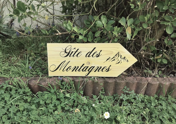 Panneau fléché extérieur en bois brut indiquant "Gîtes des Montagnes" peint en noir avec un dessin illustratif