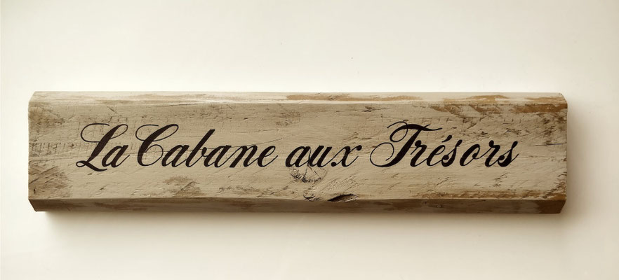 Pancarte en bois accrochée à un mur blanc, texte calligraphié en noir indiquant "La cabane aux trésors"