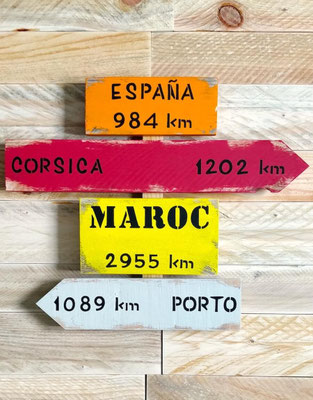 Panneau indicateur en bois recyclé et coloré, personnalisé avec des destinations comme Porto, Corsica ou Maroc, posé sur un fond bois clair