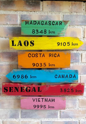 Panneau de direction à suspendre inspiration bois flotté, peinture multicolore et texte noir personnalisé avec des inscriptions géographiques comme Sénégal, Laos ou Vietnam
