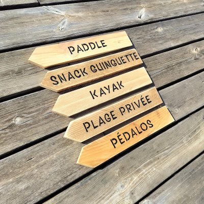 Cinq flèches en bois brut accrochées sur une palissade en bois grisé indiquant "Pédalos", "Guinguette", "Plage", ou "Paddle"